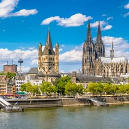 Kölner Skyline mit dem gotischen Kölner Dom und historischen Kirchen am Rhein. Bunte Gebäude säumen das Ufer unter blauem Himmel.