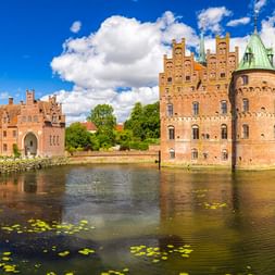 Schloss Egeskov in Dänemark, ein Wasserschloss aus rotem Backstein mit Türmen und grünen Kupferdächern, umgeben von einem Wassergraben.