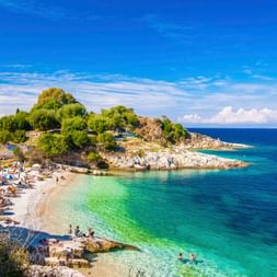 Malerischer Strand bei Lakones, Korfu mit türkisfarbenem Wasser, felsiger Halbinsel mit grünen Bäumen, Sonnenbadende am Kieselstrand unter blauem Himmel.