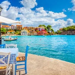 Türkisfarbene Hafenbucht mit bunten mediterranen Häusern am Hang. Blaue und weiße Stühle mit karierter Tischdecke am Ufer-Café.