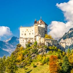 Weißes Schloss Tarasp auf grünem Hügel mit Herbstlaub, umgeben von dramatischen Schweizer Alpengipfeln unter blauem Himmel mit Wolken.
