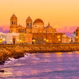 Kathedrale von Cádiz und historische Gebäude entlang der Uferpromenade bei Sonnenuntergang, mit Steinmole und lila getöntem Meer im Vordergrund.