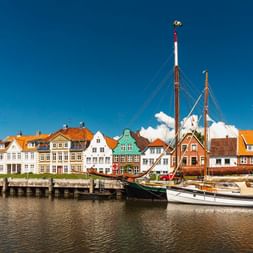 Historischer Hafen von Glückstadt mit traditionellen Segelschiffen vor bunten Giebelhäusern entlang der Uferpromenade unter blauem Himmel.