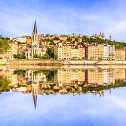 Kirche Saint-Georges mit hohem Turm und bunte Gebäude am Ufer der Saône in Lyon, perfekt im ruhigen Wasser gespiegelt.