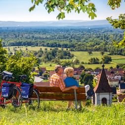 Zwei Radfahrer sitzen auf einer Holzbank mit einem roten Fahrrad daneben und blicken auf das Rheintal bei Istein mit Hügeln und Dorf.