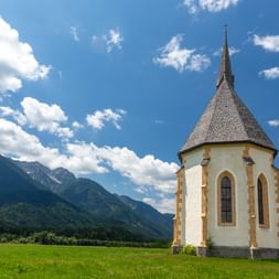 Weiße Kirche mit spitzem Turm und gotischen Fenstern in Berg im Drautal, umgeben von grüner Wiese mit Bergkette im Hintergrund unter blauem Himmel.