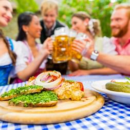 Traditionelle bayerische Brotzeit auf Holzbrett mit Obatzda. Menschen in Dirndl und Lederhosen stoßen im Hintergrund mit Bierkrügen an.