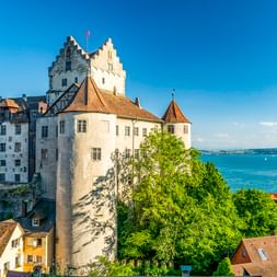 Historische Burg Meersburg mit weißen Mauern und Turm mit Blick auf den Bodensee. Häuser mit roten Dächern im Vordergrund, blauer See und Himmel.