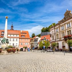 Kopfsteinpflaster-Marktplatz in Kulmbach mit bunten historischen Gebäuden, weißer Denkmalsäule und Blumenkübeln unter blauem Himmel.