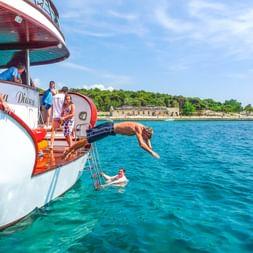 Person springt vom Boot ins türkisfarbene Wasser nahe der Insel Krk. Passagiere beobachten vom Deck, Küstenbefestigungen im Hintergrund.
