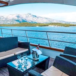 Außendeck der MS Olmissum mit schwarzen Rattanmöbeln, Weingläsern auf dem Tisch, Blick auf blaues Wasser, grüne Insel und schneebedeckte Berge.