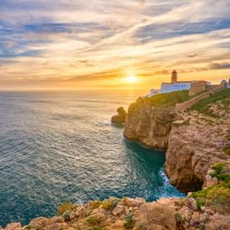 Leuchtturm Cabo de São Vicente auf dramatischen Klippen bei Sonnenuntergang. Felsige Küste mit türkisfarbenem Wasser und goldenem Himmel über dem Atlantik.