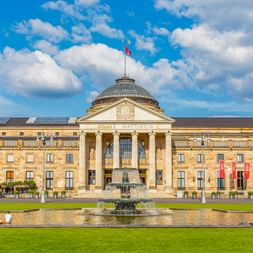 Neoklassizistisches Kurhaus in Wiesbaden mit Säulenportikus und Kuppel, mit Brunnen im Vordergrund und grüner Rasenfläche unter blauem Himmel.