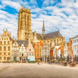 Der Turm der Kathedrale von Mechelen überragt historische Gebäude rund um einen sonnigen Marktplatz mit Straßencafés und Radfahrer.
