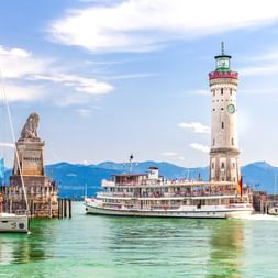 Lindauer Hafeneinfahrt mit weißem Leuchtturm, bayerischem Löwendenkmal und Raddampfer am Bodensee mit Alpenpanorama im Hintergrund.