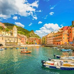 Colorful buildings surround the small harbor of Vernazza in Cinque Terre with fishing boats moored in clear water under a blue sky.