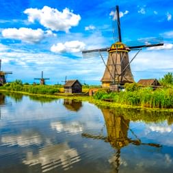 Traditionelle niederländische Windmühlen in Kinderdijk spiegeln sich in einem ruhigen Kanal unter blauem Himmel mit weißen Wolken und grüner Vegetation.
