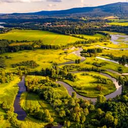 Luftaufnahme eines mäandernden Flusses durch grüne Wiesen und Wälder in Südböhmen, mit Hügeln im Hintergrund unter bewölktem Himmel.