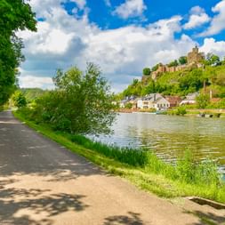 Asphaltierter Radweg entlang der Saar in Saarburg mit Burgruine auf dem Hügel, weißen Häusern am Flussufer und blauem Himmel mit Wolken.