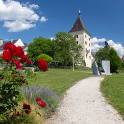 Kiesweg zum Stiftsmuseum Millstatt mit roten Rosen im Vordergrund, historischen Turmgebäuden und grünen Rasenflächen unter blauem Himmel.