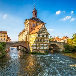 Historisches Fachwerkgebäude des Alten Rathauses von Bamberg auf Insel in der Regnitz, verbunden durch steinerne Brückenbögen unter blauem Himmel.