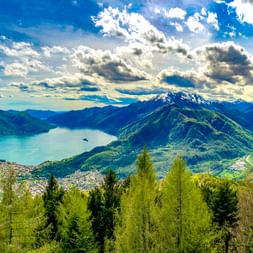 Panoramablick auf den Lago Maggiore, umgeben von grünen Bergen unter dramatischem Wolkenhimmel. Bewaldete Hänge rahmen den türkisfarbenen See.