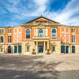 Das Bayreuther Opernhaus mit seiner markanten orangeroten Fassade und cremefarbenen Ornamenten unter blauem Himmel mit weißen Wolken.