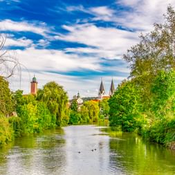 Fluss Altmühl fließt durch üppiges Grün in Richtung Eichstätt mit Kirchtürmen und rotem Uhrturm vor blauem Himmel.
