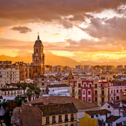 Málaga Stadtbild bei Sonnenuntergang mit Kirchturm über den Gebäuden. Orange-goldener Himmel mit Bergen im Hintergrund.