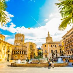 Sonniger Platz in Valencia mit Brunnen, Palmen und historischen Gebäuden, darunter die Kathedrale von Valencia mit Glockenturm unter blauem Himmel.