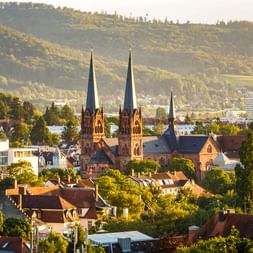 Johanneskirche mit zwei Türmen in Freiburgs Stadtbild, umgeben von roten Ziegeldächern und grünen Bäumen, mit bewaldeten Hügeln im Hintergrund.