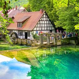 Kristallklarer türkisfarbener Blautopf in Blaubeuren mit Fachwerkhaus und hölzerner Aussichtsplattform, umgeben von grünen Bäumen.
