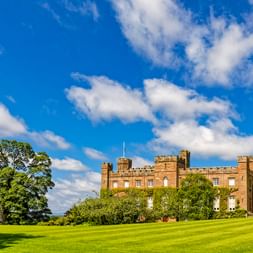 Scone Palace bei Perth mit zinnenbesetzten Türmen und gotischen Fenstern, umgeben von grünem Rasen und alten Bäumen unter blauem Himmel.