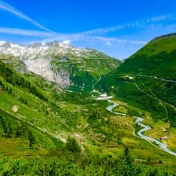 Grimselpass-Tal mit mäanderndem Fluss und Serpentinenstraße durch grüne Almwiesen, umgeben von steilen Bergen unter blauem Himmel.