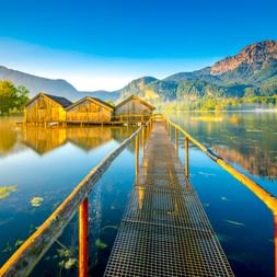 Metallsteg führt zu Bootshäusern am ruhigen Kochelsee mit Bergspiegelung. Bayerische Alpen und bewaldete Hänge unter blauem Himmel.