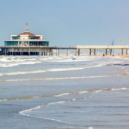 Sandstrand in Blankenberge mit Wellen am Ufer. Ein rundes Seebrückenpavillon und eine lange Seebrücke erstrecken sich in die Nordsee unter blauem Himmel.