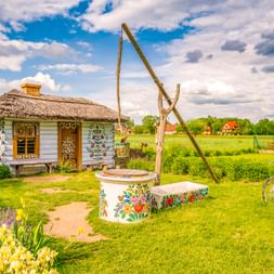 Traditionelles Reetdachhaus mit Blumenmalereien in Zalipie. Ein verzierter Brunnen mit Holzrahmen steht im Garten, ein Fahrrad daneben.