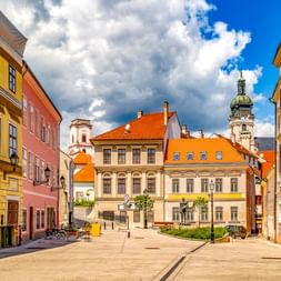 Bunte Barockgebäude umgeben einen Fußgängerplatz in der Altstadt von Győr, mit einem Kirchturm im Hintergrund unter blauem Himmel.