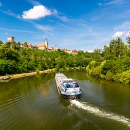 Passagierschiff auf dem Neckar mit Bad Wimpfens historischer Altstadt und Turm auf einem grünen Hügel unter blauem Himmel.