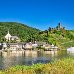 Dorf Beilstein an der Mosel mit weißer Kirche, traditionellen Häusern und Ruine Burg Metternich auf weinbergbedecktem Hang.