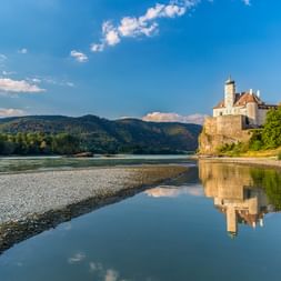 Schloss Schönbühel auf einem Felsvorsprung an der Donau, mit bewaldeten Hügeln im Hintergrund und Spiegelung des Schlosses im ruhigen Wasser.