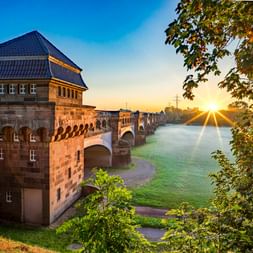 Historische Steinbrücke und Turmgebäude am Mindener Wasserstraßenkreuz bei Sonnenaufgang, mit der Weser darunter und grünem Laub im Vordergrund.