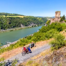 Radfahrer rasten auf Weg nahe Burg Gutenfels mit Blick auf den Rhein bei Kaub, mit grünen Hügeln und blauem Himmel.
