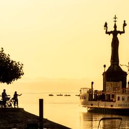 Silhouette der Imperia-Statue und Hafengebäude in Konstanz bei Sonnenaufgang. Zwei Radfahrer ruhen unter einem Baum am Ufer des Bodensees in goldenem Licht.