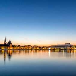 Aarhus Hafen bei Sonnenuntergang mit beleuchteten Gebäuden, die sich im ruhigen Wasser spiegeln. Kirchturm links, moderne Gebäude rechts.
