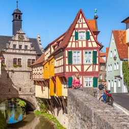 Radfahrerin auf Steinbrücke in Marktbreiter Altstadt mit bunten Fachwerkhäusern, historischem Torturm und kleinem Kanal darunter.