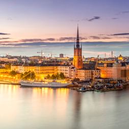 Stockholmer Uferpromenade bei Sonnenuntergang mit beleuchteten historischen Gebäuden, die sich im ruhigen Wasser spiegeln. Kirchturm ragt über die Stadtlandschaft unter lila Himmel.