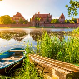 Marienburg mit roten Backsteintürmen und Mauern am ruhigen Fluss. Ein blaues Boot und eine Holzbank liegen zwischen Schilf im Vordergrund.