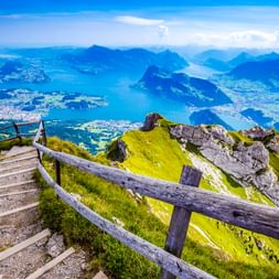 Holzstufen und Zaun auf dem Pilatus mit grünen Hängen und Felsvorsprüngen. Vierwaldstättersee und umliegende Berge unter blauem Himmel sichtbar.
