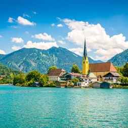 Bayerisches Dorf am Tegernsee mit Kirchturm, traditionellen Gebäuden und bewaldeten Bergen unter blauem Himmel mit weißen Wolken.